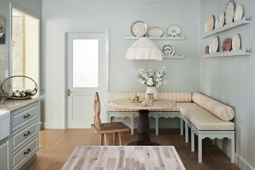 pale blue-green kitchen nook with vintage plates on wall shelves, custom upholstered striped bench seat with stone table and a large ruffle linen pendant light hanging above